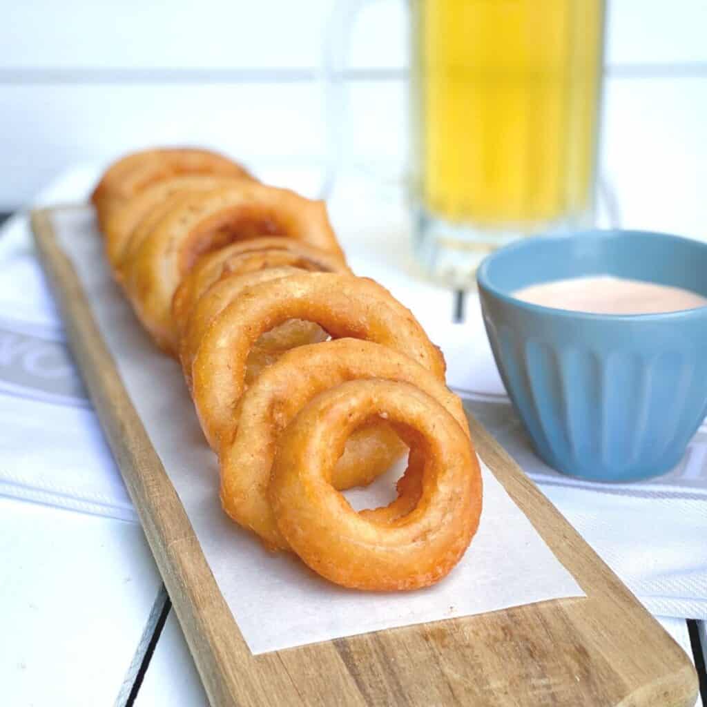 crispy beer battered onion rings on a wooden plank with dipping sauce and a beer on the side