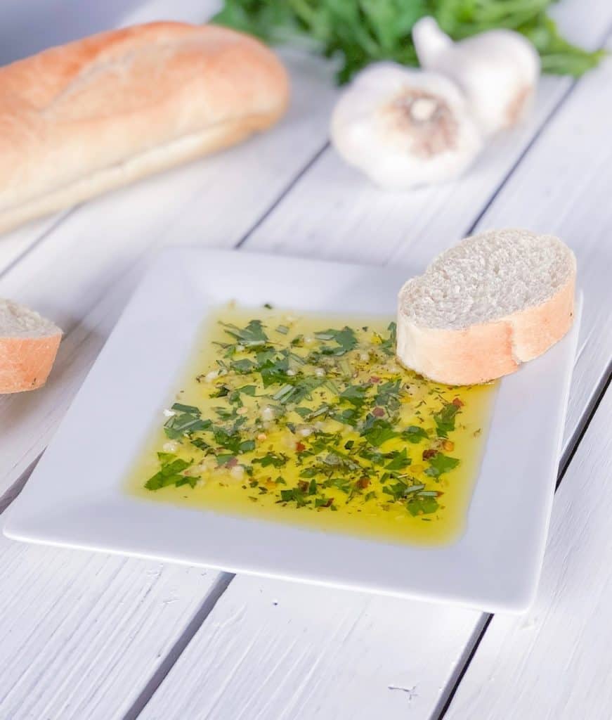 herbs in olive oil on a white plate with a slice of bread next to it