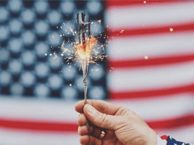 A lit sparkler with the American flag in the background.