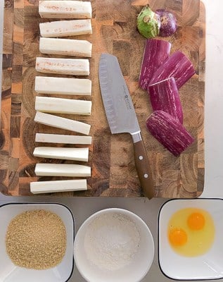 Eggplant fries on a board with breadcrumbs, flour and eggs.