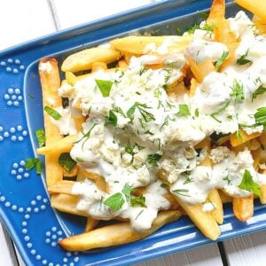 Greek french fries covered with feta cheese and yogurt sauce on a blue plate.