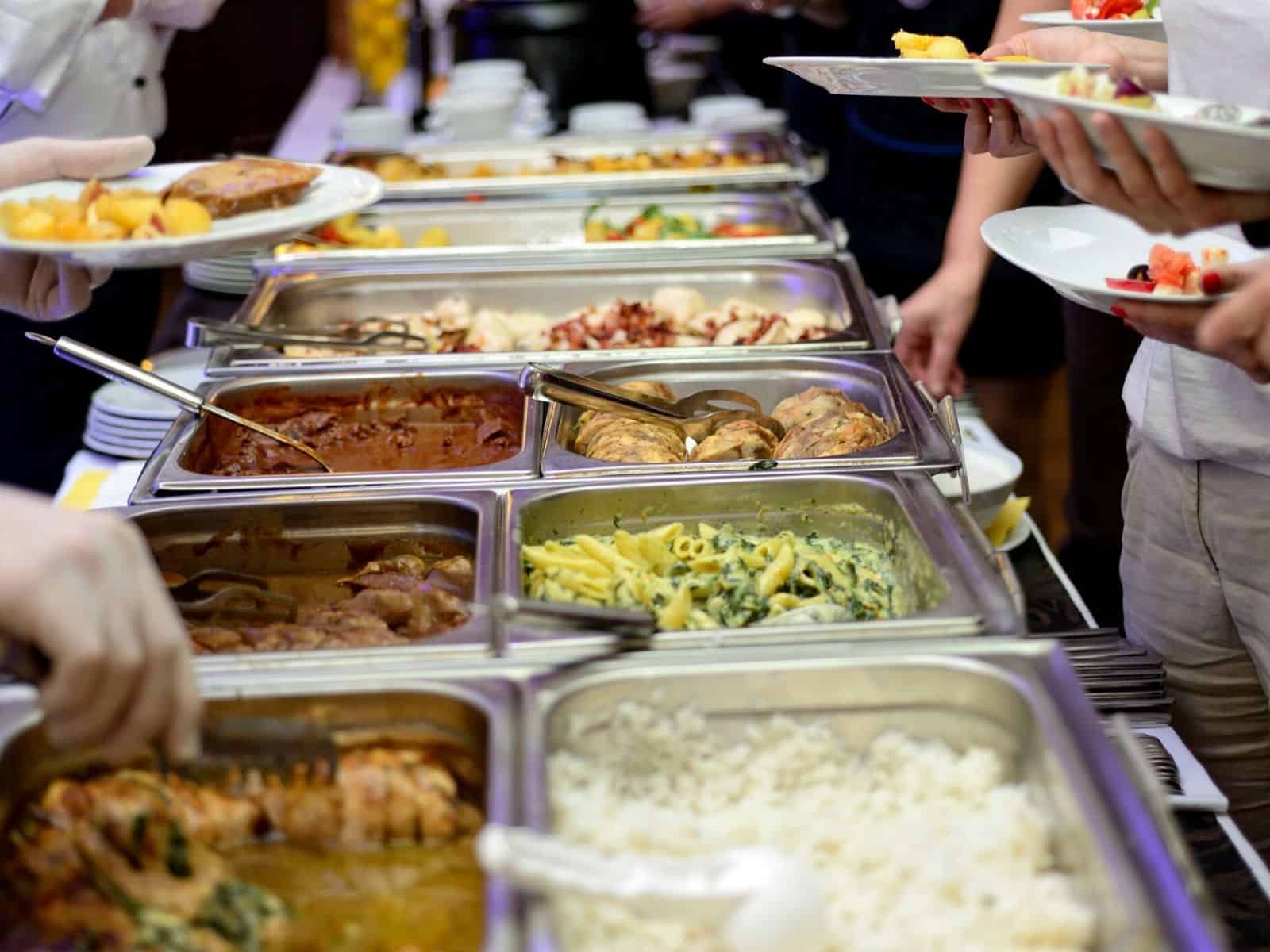 chafing dishes set up on a buffet table with warm food and people serving themselves