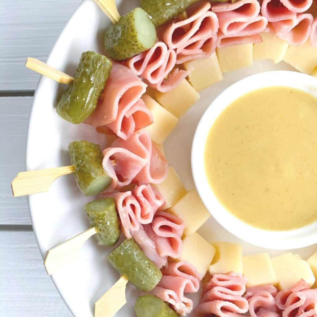 skewered ham cheese and pickle appetizer on a plate with dipping sauce in center.