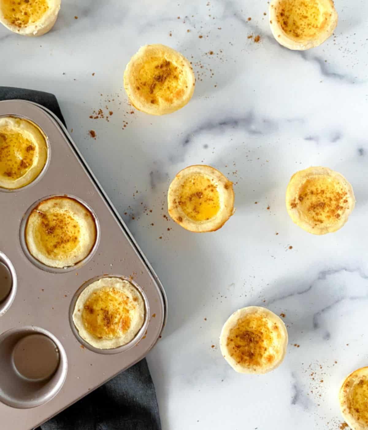 Mini egg custard tartlets in a muffin tin sprinkled with nutmeg on table.