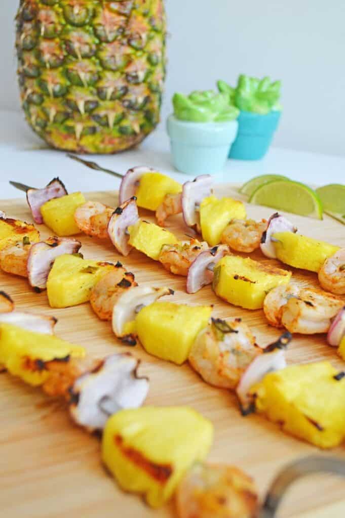 Cooked skewers of pineapple, chicken and onion on wooden cutting board with a garnish of lime slices. 