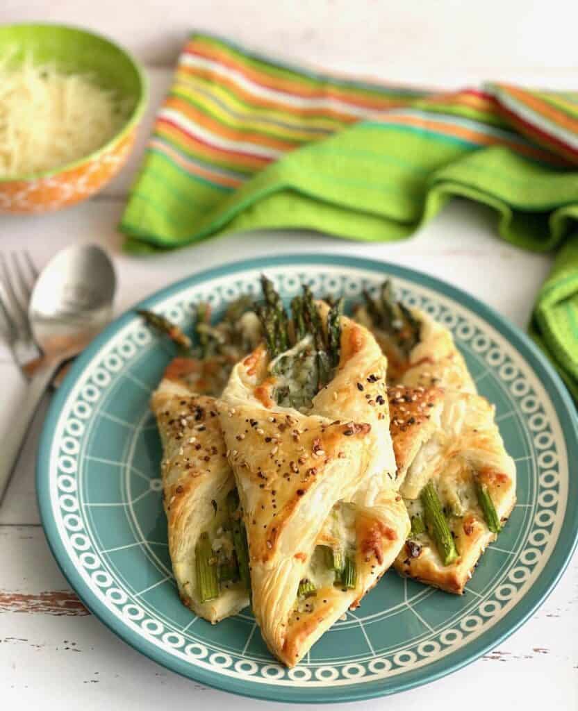 Puff pastry folded around asparagus spears with sesame seeds on top sitting on a blue patterned plate with a fork and spoon sitting next to it.