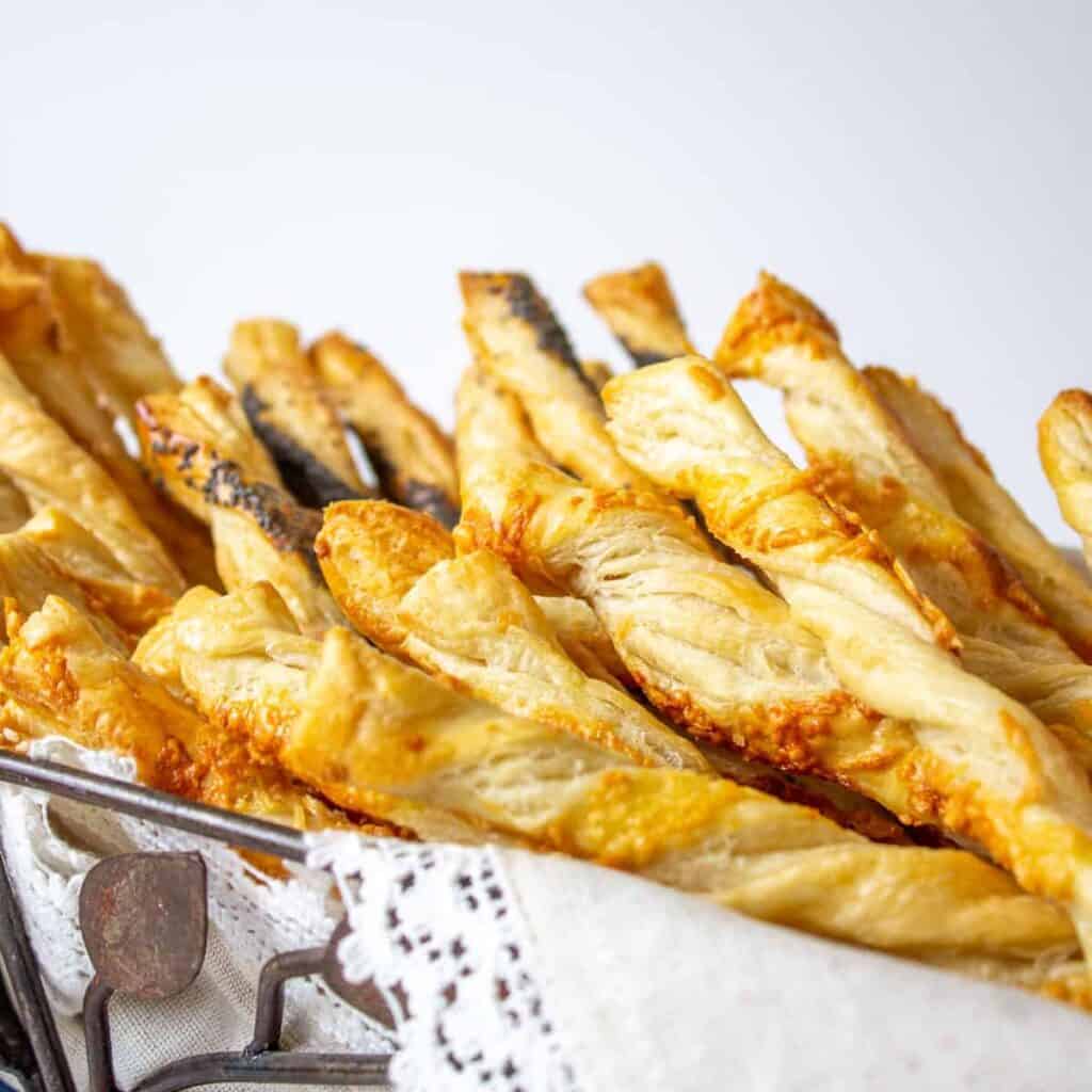 A pile of cheese straws in a wire basket lined with a white linen.