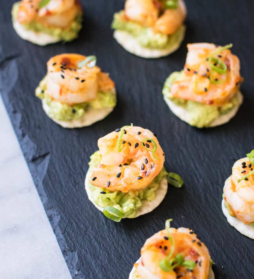 Crackers with avocado cream, shrimp, and black sesame seeds sitting on a black cutting board. 