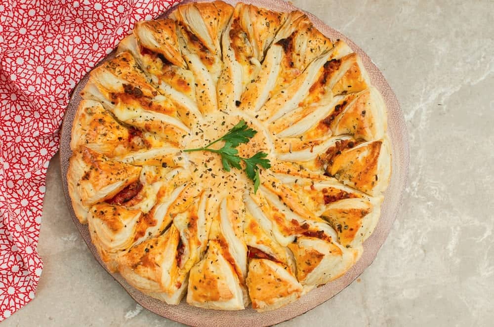 A puff pastry pizza on round tray on a marble counter with a red print cloth behind it.