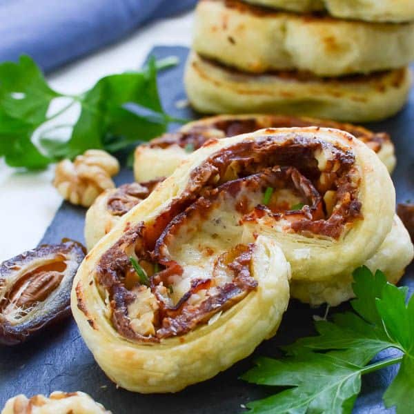A close up of a prosciutto and date palmier and laying on a stack of them and green leaf garnish with a half of a date on a black cutting board.