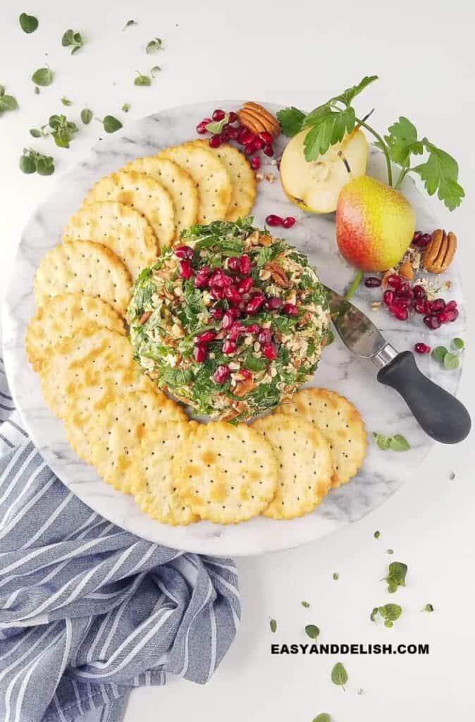 Cheese ball rolled in nuts and green garnish with pomegranate seeds on top and crackers layered around it and a pear.