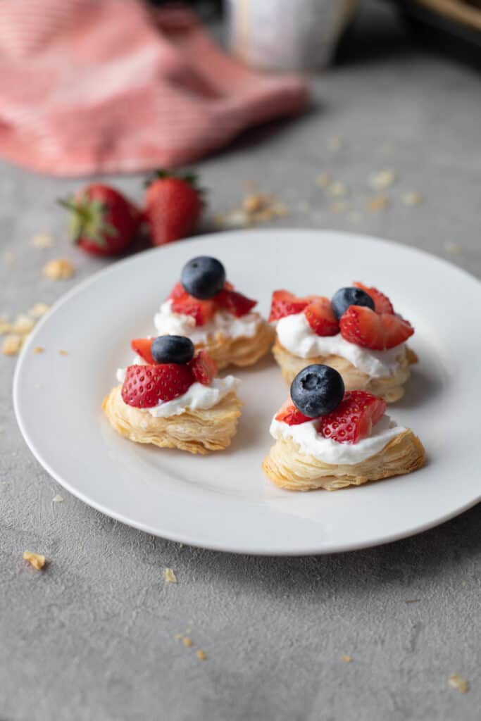 Puff pastry bites with whipped cream and fruit on top laying on round white plate with strawberries in the background.
