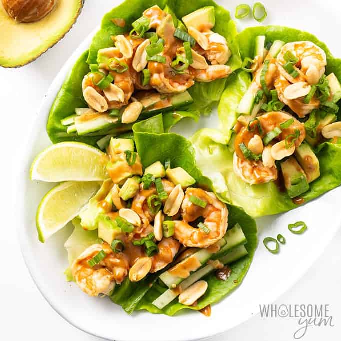 3 lettuce wraps with shrimp filling and peanuts on top laying on a white plate with lemon wedges.