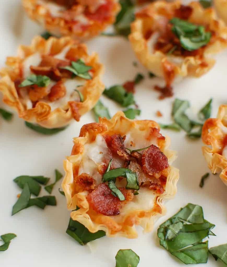 Close up of a bacon pizza bite surrounded by chopped basil.
