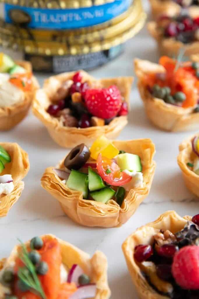 Phyllo cups filled with avocado, cherry tomatoes, cucumber, hummus, and a black olive. 