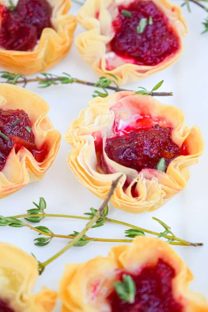 A close up of a phyllo cup filled with melted brie with cranberry sauce on top with fresh thyme laying next to it. 