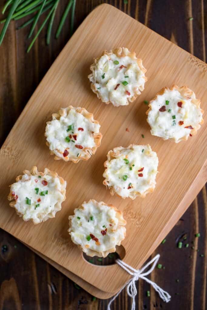 Six phyllo cups filled with artichoke dip with a red pepper flakes and green onion garnish sitting on a wooden cutting board. 