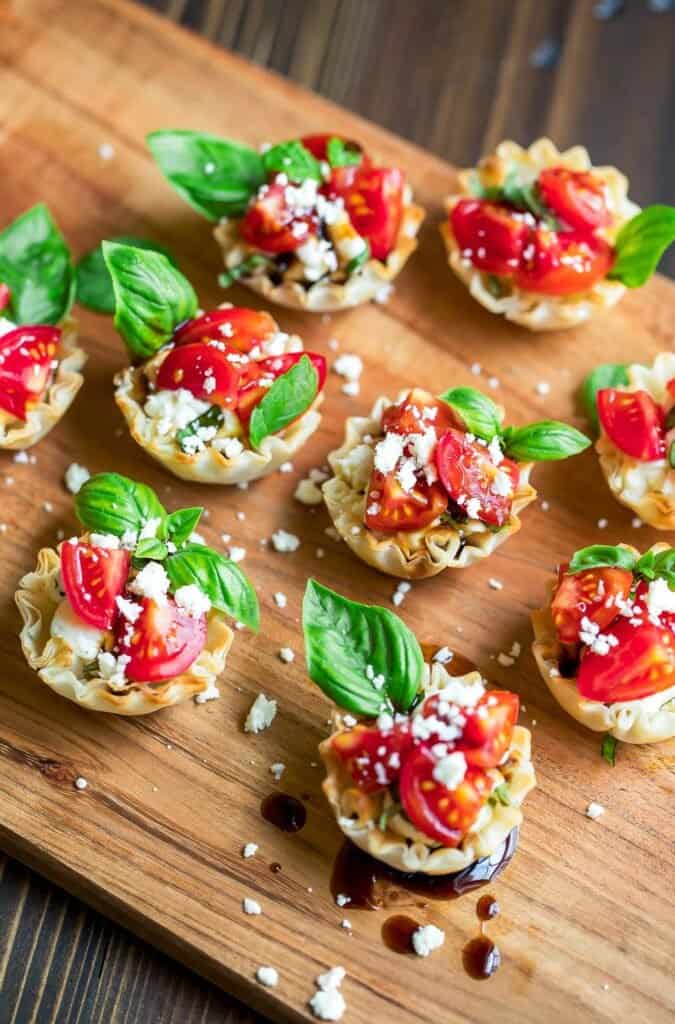 Rows of bruschetta phyllo cups with whole basil leaves and feta cheese sprinkled on top sitting on a wooden surface.