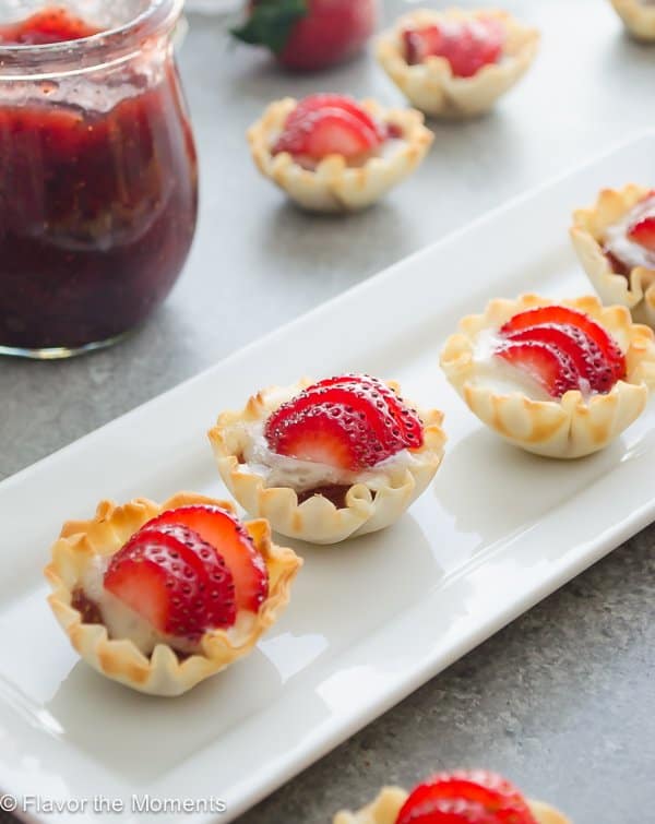 A row of phyllo cups filled with melted brie and topped with a fresh sliced strawberry on rectangular plate with more bites and a jar of jam in the background.
