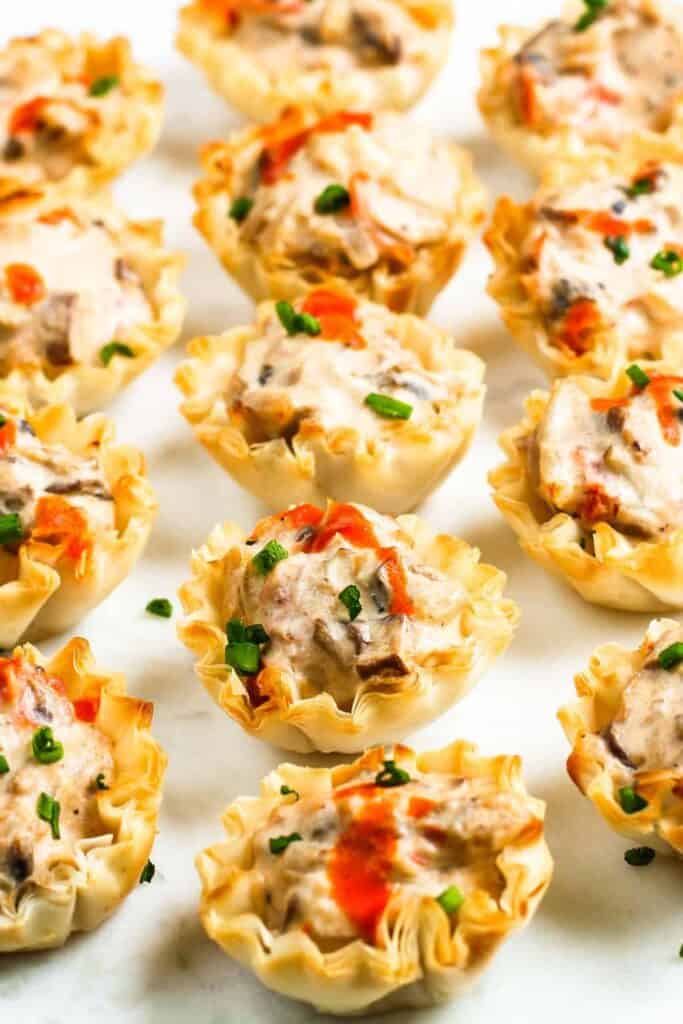 Rows of Creamy Mushroom Tartlets on a white surface with hot suace and a chive garnish.
