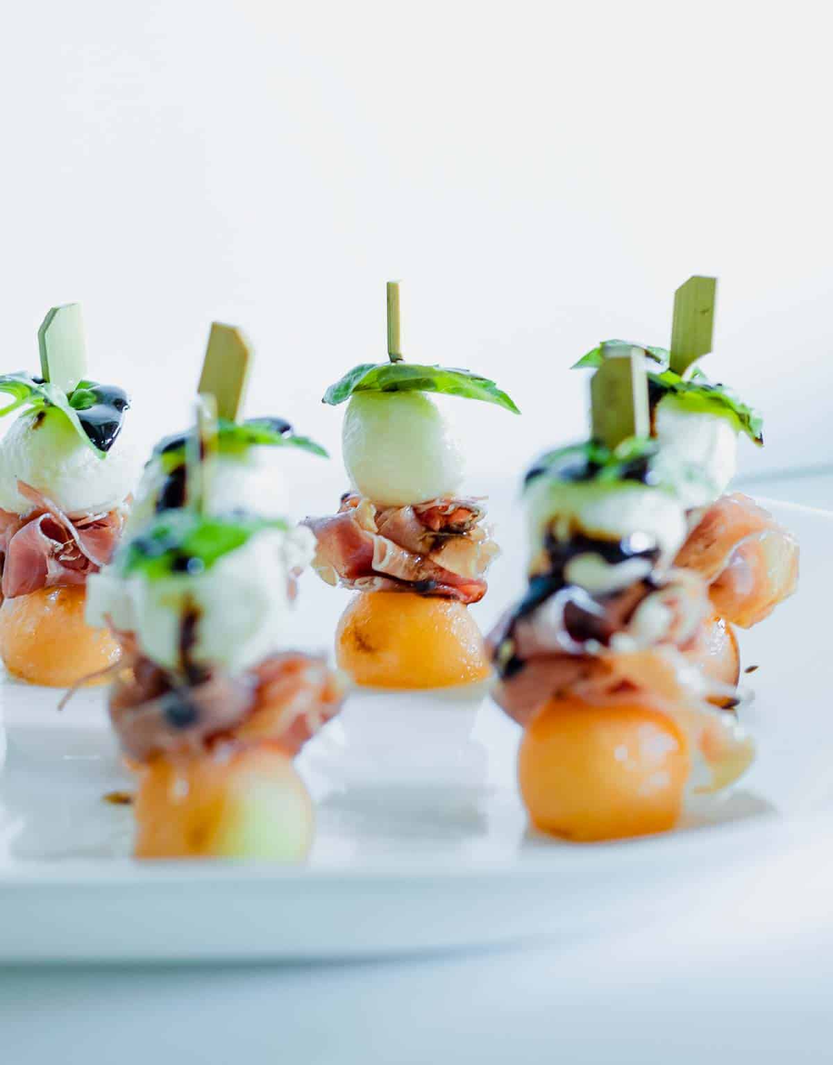 Prosciutto, melon, mozzarella, basil skewers on a plate with balsamic glaze.