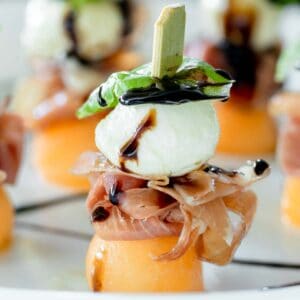 Prosciutto melon mozzarella skewers on a plate drizzled with balsamic glaze.