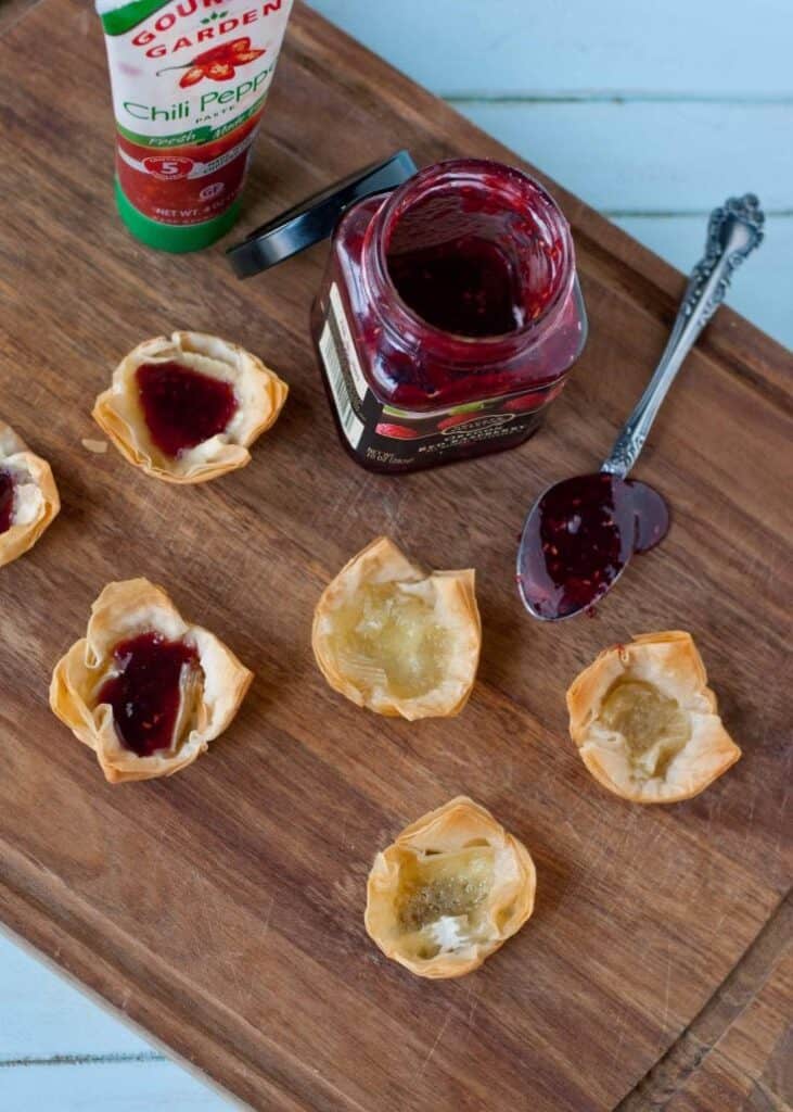 Two filled phyllo cups and three empty ones on a wooden cutting board with a jam jar and a spoonful of jam laying next to them. 