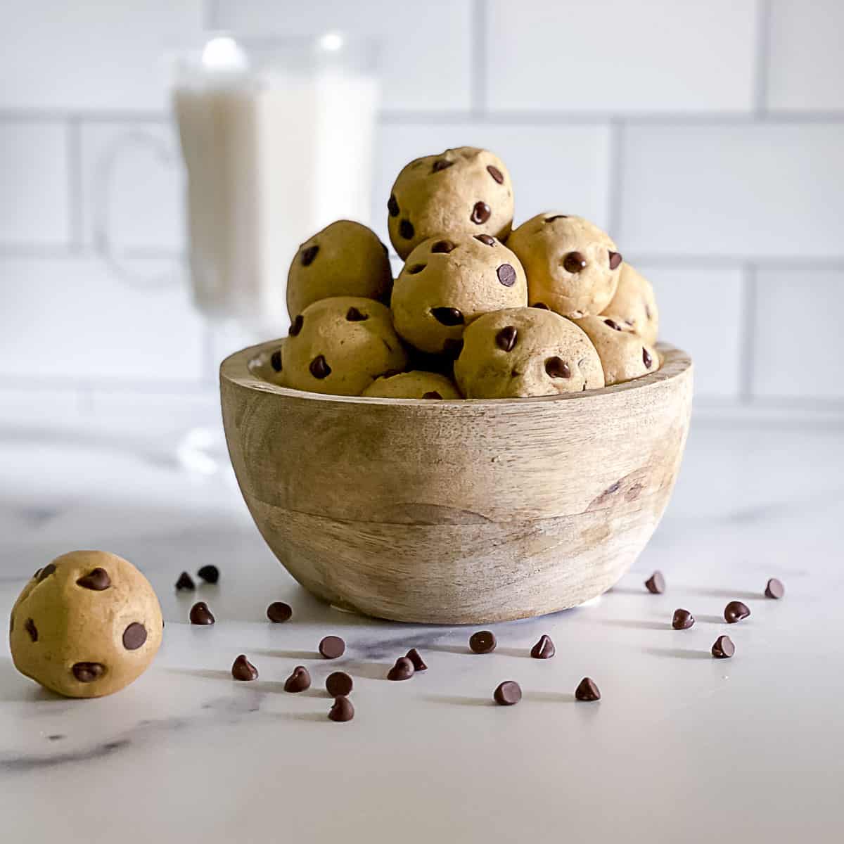 Edible no bake chocolate chip cookie dough balls stacked in a bowl with chips sprinkled on table.