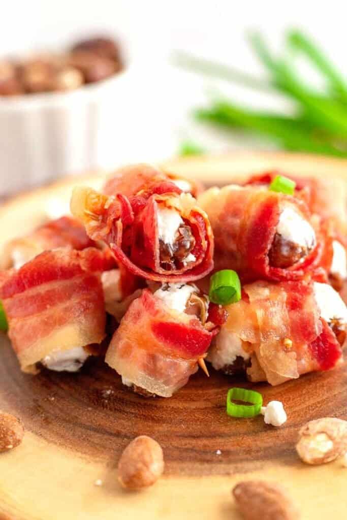 A pile of bacon-wrapped dates stuffed with goat cheese on a wood slice with green onion and almond garnish. 
