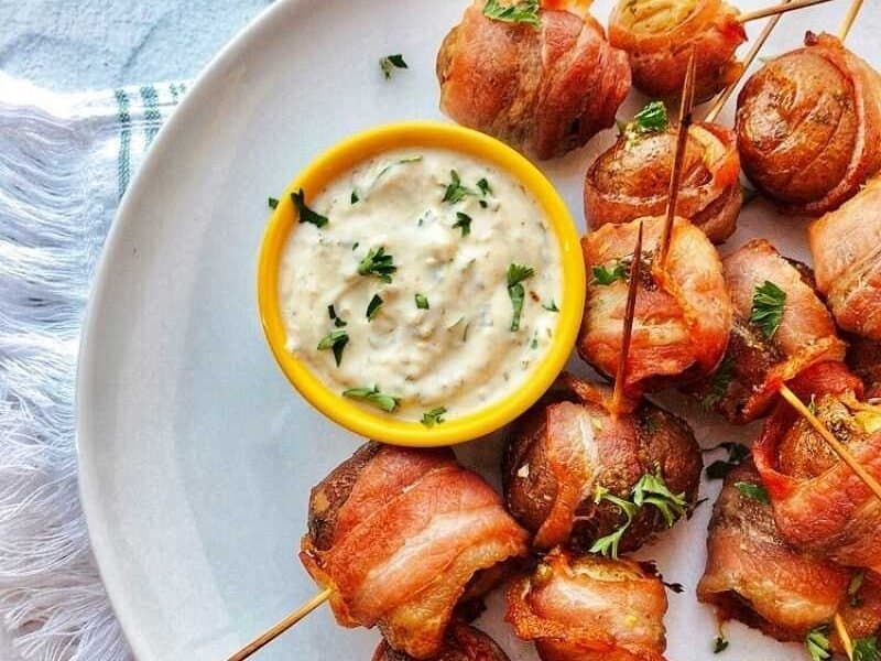 Bacon-wrapped small potatoes on a round white plate with a small yellow bowl of sauce.