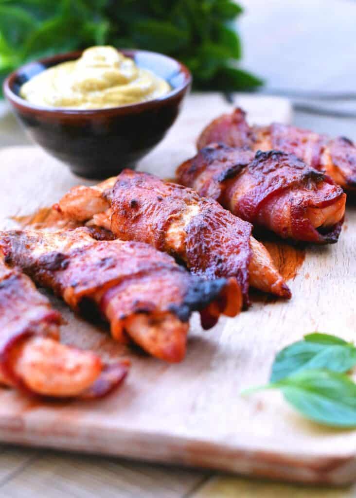 Bacon wrapped chicken strips laying on a wood surface with a bowl of dipping sauce sitting next to them.