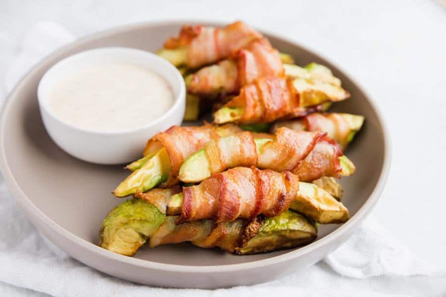 A grey plate sitting on a white towel. The plate is full of bacon wrapped avocado with a small bowl of dip sitting next to it. 