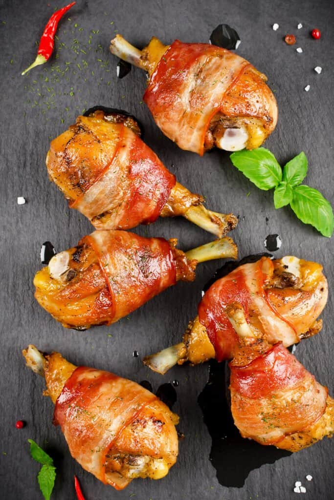 Bacon wrapped chicken legs on a black surface with green leaf garnish.
