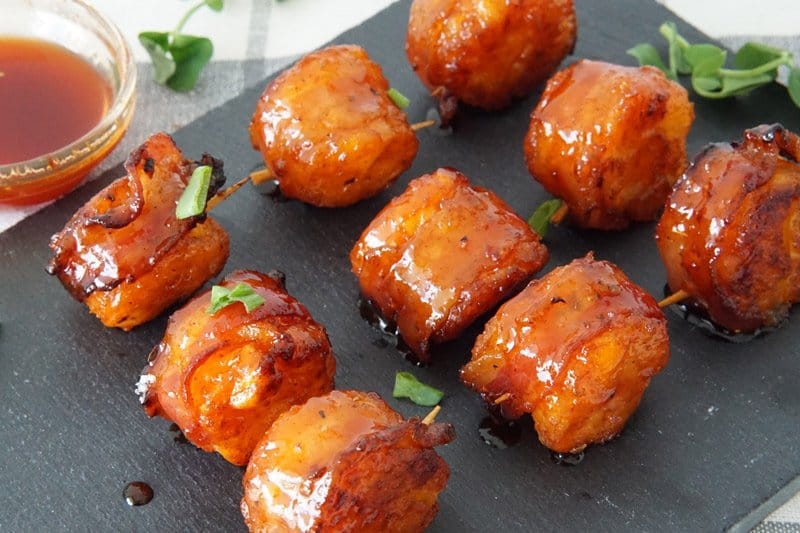 Bacon wrapped tater tots with sriracha honey glaze on a black cutting board with a green leaf garnish and a small bowl of sauce next to them.