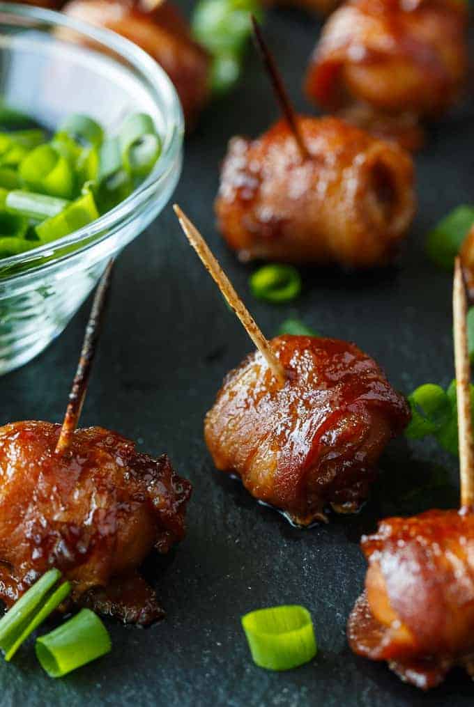 Bacon-wrapped water chestnuts with toothpicks and a green onion garnish on a black surface next to a clear bowl full of sliced green onions.