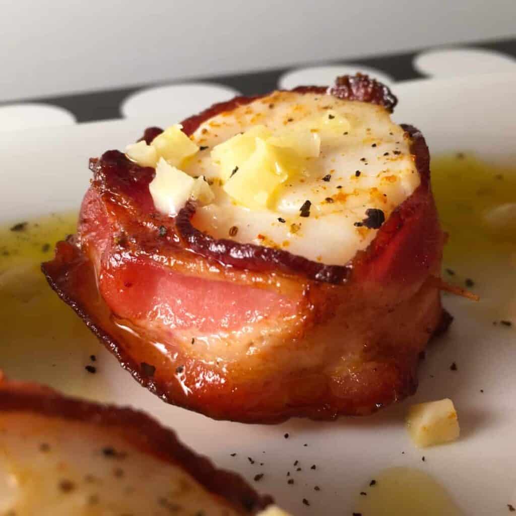 A close up of a bacon wrapped scallop on a white surface drizzled with garlic butter.