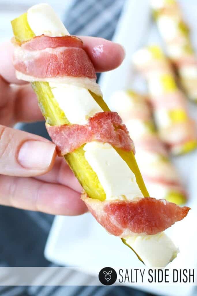A hand holding a bacon-wrapped pickle stuffed with cream cheese.