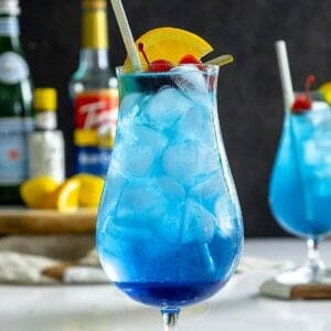 Blue lagoon mocktail recipe on table with cherry and orange slice.