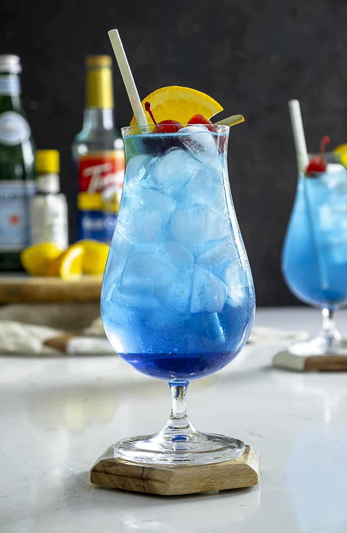 Blue lagoon mocktail recipe on coaster with orange and cherry on top.