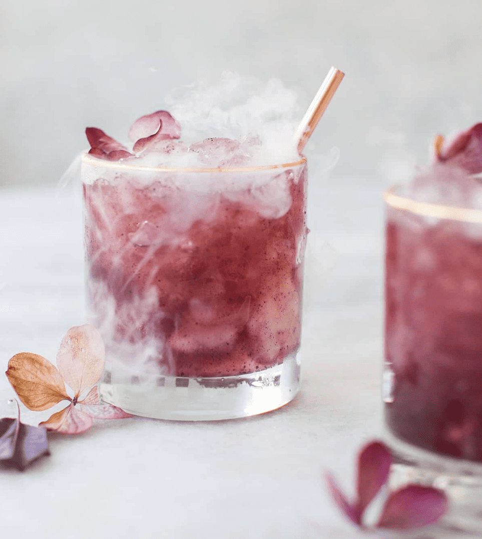 Purple cocktails on a table with flowers and smoke.