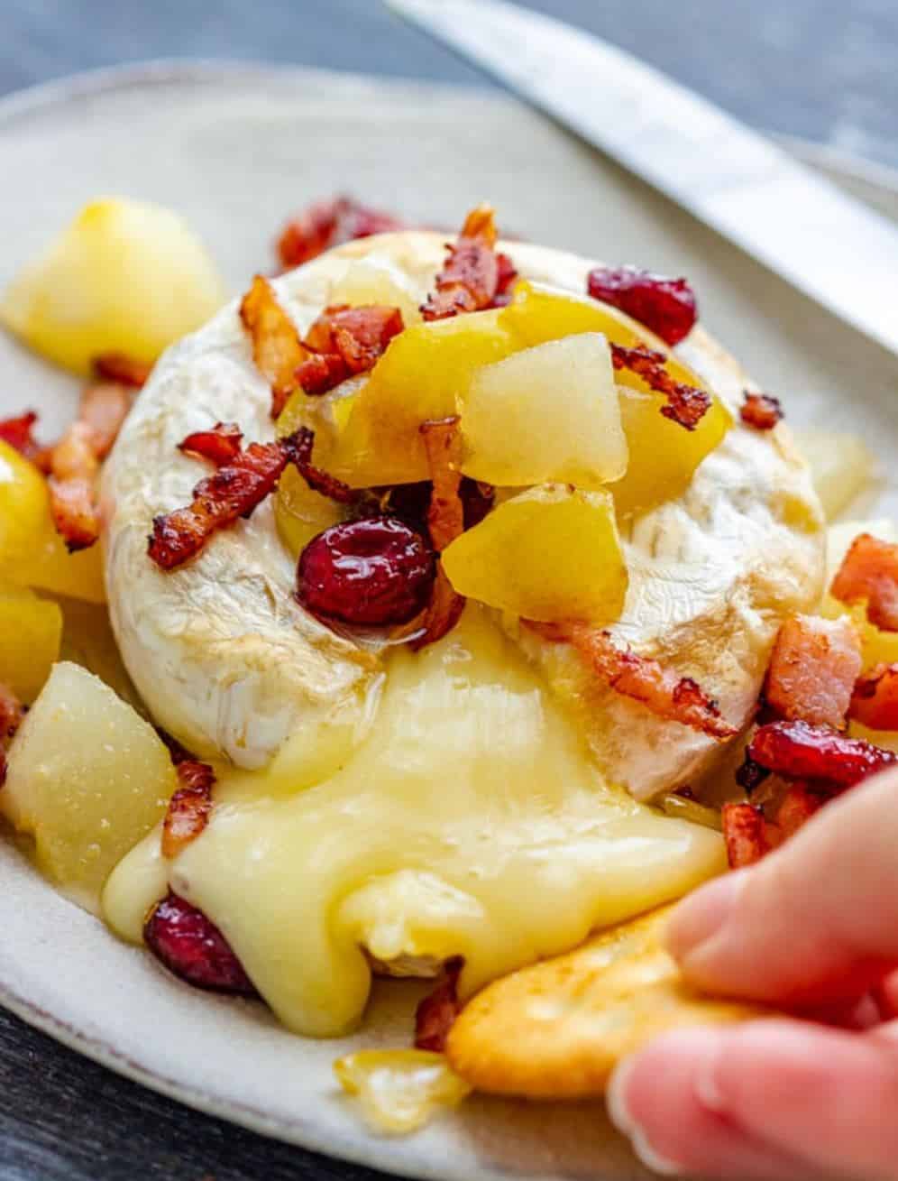 Baked brie with pear and bacon with a hang dipping a cracker.