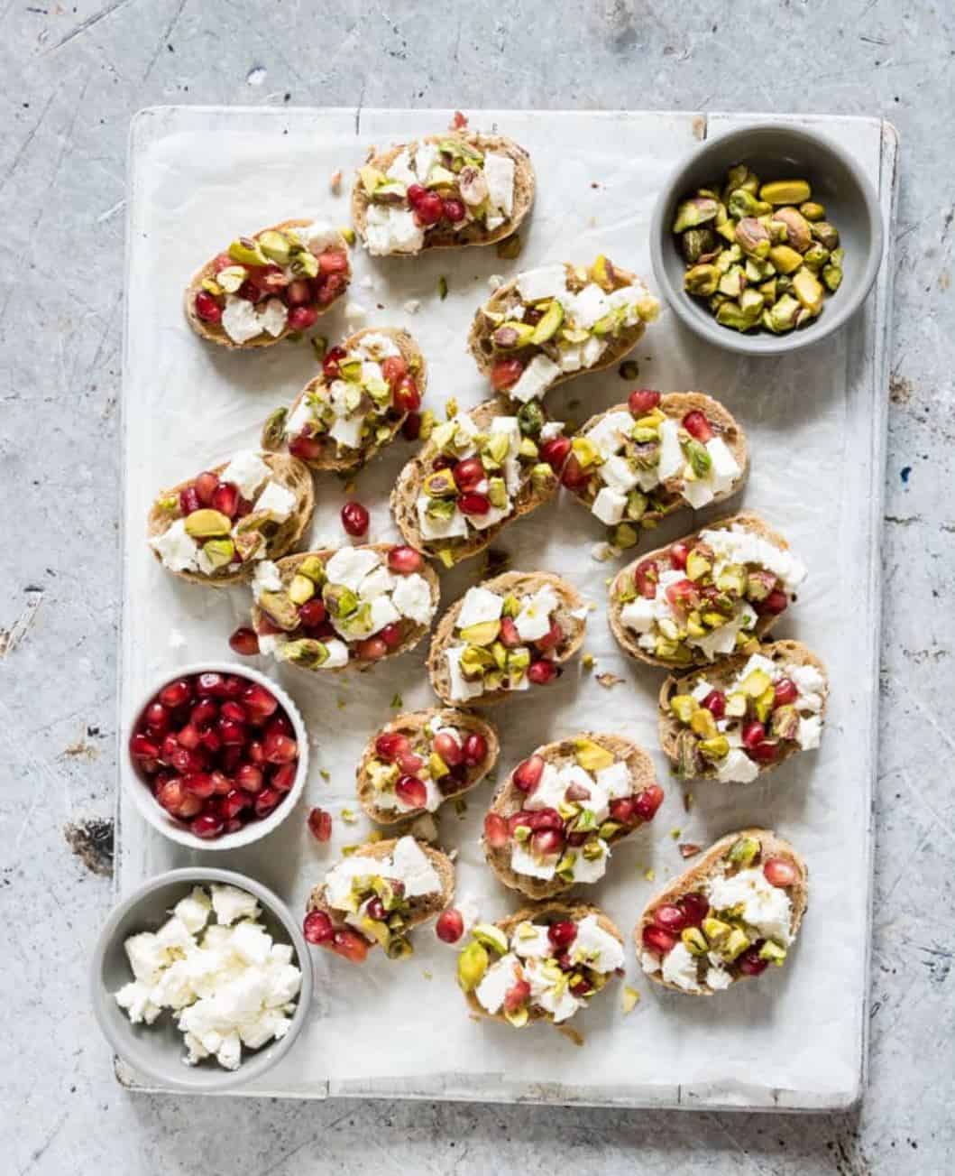 Crostinis with pomegrant, cheese and pistachios on a white serving board. 