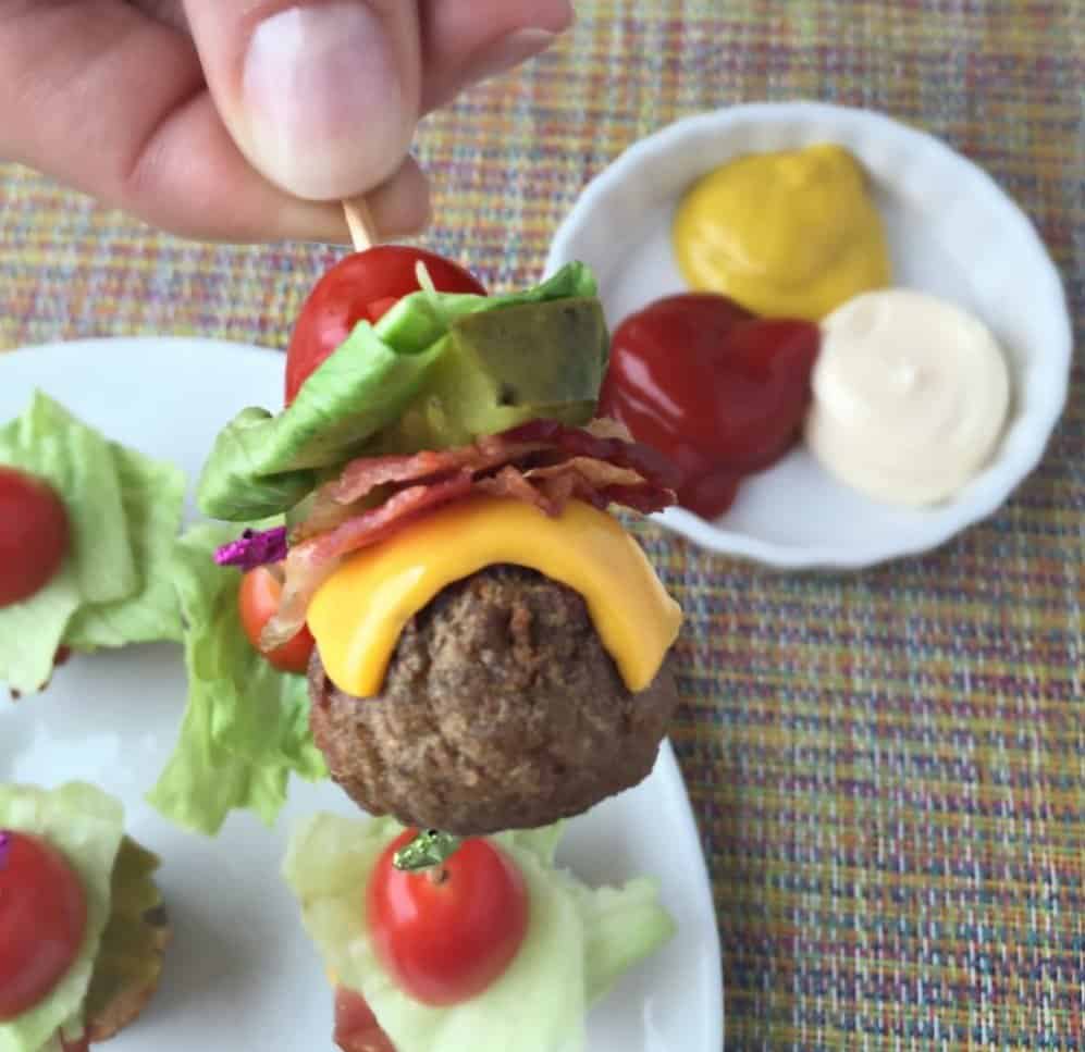 A meatball with hamburger fixings on a skewer. With dipping sauce in the background. 