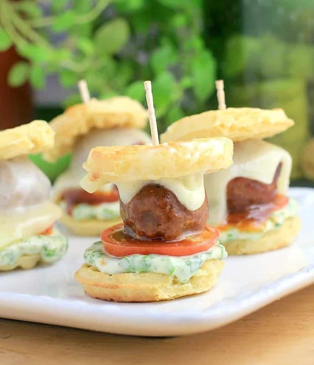 Meatball sliders with melted cheese and other fixings, on a white serving plate. 