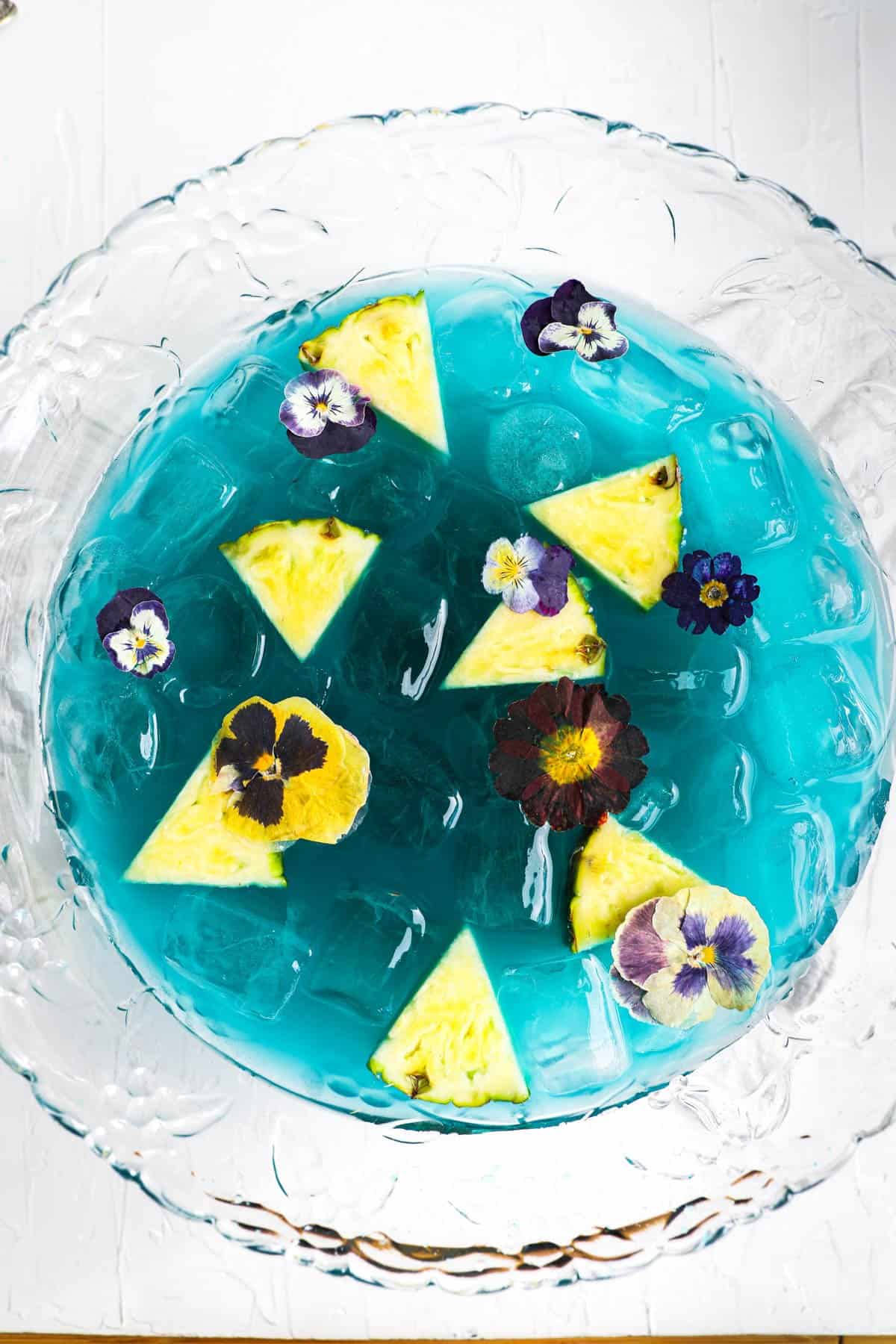 Blue alcoholic baby shower punch in bowl with pineapple chunks and flowers.