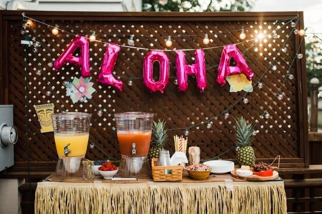 Tropical party theme table with "Aloha" backdrop and food and drinks on a table.