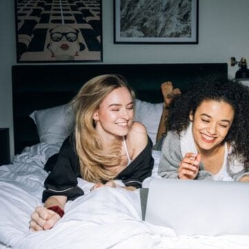 Two women laying on a bed laughing at a laptop.