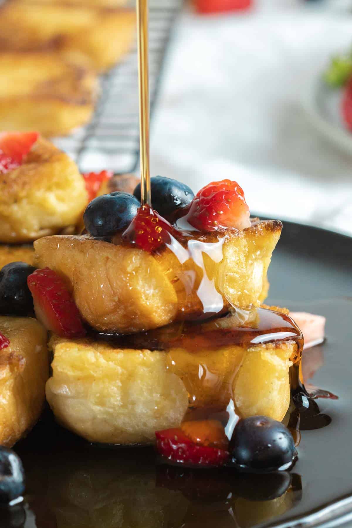 Close up of Hawaiian roll french toast with berries and maple syrup on a dark plate.