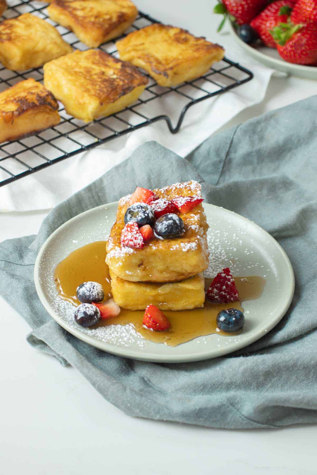 Hawaiian roll french toast stacked on a small plate with syrup and fruit.
