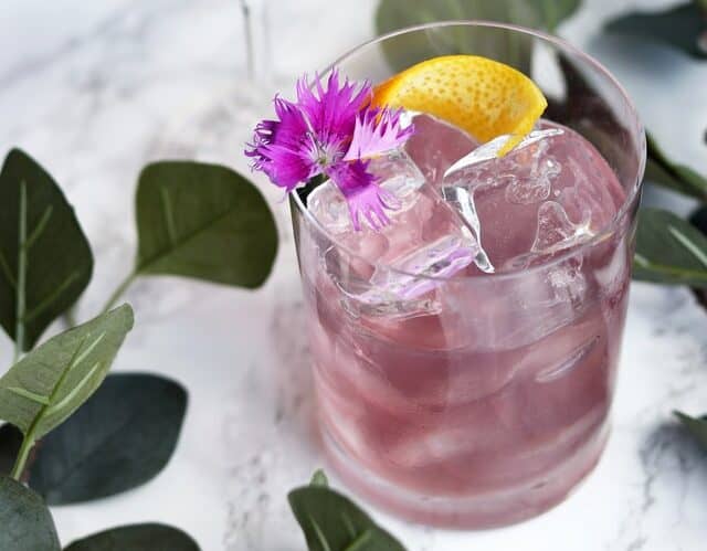 A purple cocktail garnished with a flower and lemon on a white marble table.